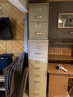 Pair of older metal filing cabinets. One tall five-drawer and one shorter two-drawer, placed side by side against a wall near a wooden desk and cardboard boxes.