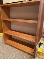 Wooden shelving unit with three shelves, medium-tone wood with natural grain and knots, standing on carpet