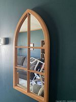 Frontal view of the wooden framed arch-top mirror hanging on a dark green wall showing its six-pane divided mirror surface and light wood frame.