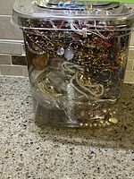 Clear plastic jar filled with various vintage jewelry including chains, cameo, and pendants, placed on a kitchen counter.