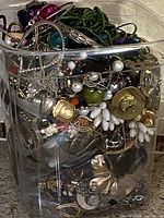 Jar filled with mixed vintage jewelry showing various styles of necklaces, bracelets, and other jewelry packed inside.
