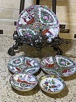 Five Asian ceramic dishes displayed on counter; four with segmented traditional figure designs in pink, green and orange, one smaller with butterfly floral pattern