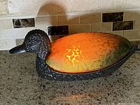 Side view of vintage mallard duck night light lamp with bronze body and amber-green art glass shade turned on, showing warm glowing light.