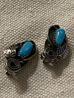 Close-up image of both earrings showing the turquoise stones and silver leaf and swirl designs on a textured surface.