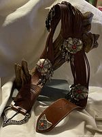 Full view of the brown leather belt with large engraved metal conchos featuring red stones, displayed hanging on wood.