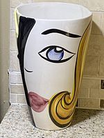 Front view of white ceramic vase with painted mid-century modern woman's face including blue eye, black eyebrow, pink lips, and yellow hair.