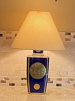 Blue porcelain table lamp with gold decorative emblems and beige textured lampshade, photographed against kitchen backsplash.