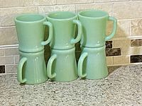Photo showing six vintage pale green opaque glass mugs with D handles stacked in two rows.