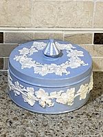 Front view of vintage Wedgwood round trinket box showing lid and embossed cream floral decoration on blue ground.