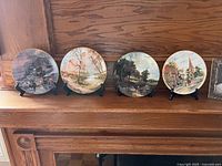 Lot of 4 collector plates displayed side by side on wooden mantel with black stands.