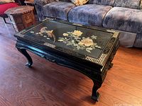 Front angled view of black lacquered coffee table with glass top showing birds and floral Chinoiserie painting.