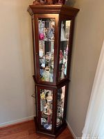 Side view of tall two-tier curio cabinet showing wood frame and glass panels with collectibles inside (not included in lot).