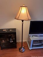 Full view of the floor lamp next to a small black cabinet and TV stand, lamp turned on showing warm light through beige fabric shade.