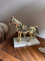 Gold metal horse sculpture standing on white marble base placed on wood surface, showing the figurine's shiny gold color with detailed saddle and mane.