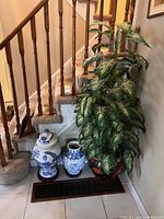 Three-piece lot showing faux plant in black and red floral planter and two decorative ceramic vases beside a staircase.