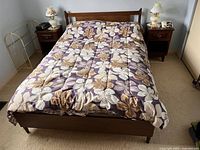 Front view of vintage queen size wooden bed frame with floral bedspread, flanked by two dark wood nightstands each topped with a lamp and telephone. Brass blanket stand visible beside left nightstand.