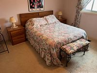 Overall setup of the bedroom lot showing vintage queen headboard, two wooden nightstands with lamps, floral bedspread, and wrought iron floral bench at foot of bed.