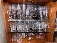Full view of three shelves inside a wooden cabinet showing assorted crystal and glassware including champagne flutes, wine glasses, highball glasses, cut crystal tumblers, brandy snifters, decanter with stopper, shot glasses, miniature glass figurine wine bottle on a cart, and small candle holder.