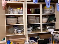 Wide view of three-section wooden kitchen cabinet showing all included items: ceramic plates, glass bowls, plastic containers, metal trays, and assorted kitchenware. Top shelves hold glassware and pitchers; middle shelves have plates and containers; bottom shelves contain serving pans and trays.