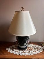 Front view of the oriental style table lamp on a wooden surface with a lace doily underneath. Shows the black ceramic base with floral design and white lampshade.