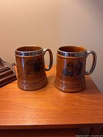 Front and side views of a pair of brown ceramic beer steins with metal rims and handles on a wooden surface.