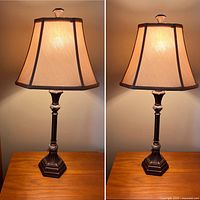 Pair of matching table lamps showing metal hexagonal bases, slender vertical stems with decorative ring, beige fabric shades with dark trim, and clear finials on top
