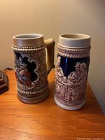 Front view of two vintage German beer steins showing detailed relief and painted designs.