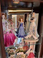 Photo showing five porcelain figurines on glass shelf inside cabinet, featuring various female figures in colorful gowns including one with geese.