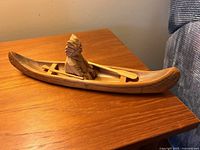 Side view of small hand-carved wooden canoe showing figure wearing feathered headdress seated inside, with paddle beside them.
