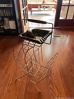 Angled full view showing the metal frame with curved design, black ashtray with ridged edges, and wire magazine rack at base.