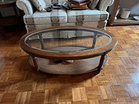 Oval vintage coffee table with glass top inset in wood frame, cane bottom shelf, placed on parquet floor in front of sofa