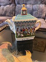Front and side views of the vintage Sadler teapot depicting a tennis match with players and spectators around a pavilion, with clock tower lid.