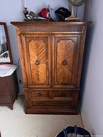 Front view of vintage wooden armoire showing the two paneled doors with chevron patterned wood grain and three bottom drawers with brass ring pull handles, placed against a wall with some hats and a fan on top.