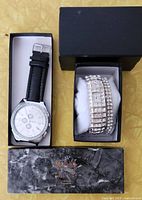 Two watches displayed: silver-tone round watch with black strap and crystal accents, and matching women's crystal bracelet watch with rectangular face in black box.