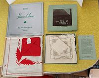 Two boxed sets of vintage table linens: one with red embroidered cream tablecloth and red napkins, the other with white linens with brown embroidery and matching napkins in box with pamphlet.