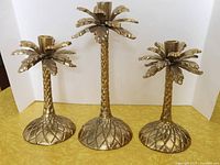 Front view of three brass palm tree candlesticks showing height differences and design details.