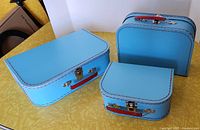 Three small vintage blue paperboard suitcases with red handles shown from the front on a yellow surface.