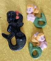 Three vintage mermaid wall decorations including a black chalkware Favor Ware piece and two ceramic mermaids with blonde hair and green tails on a yellow background.