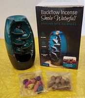 Backflow incense smoke waterfall burner with two bags of incense cones in front on yellow surface and the product box behind.