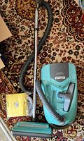 Full view of the teal Kenmore canister vacuum with hose, telescopic wand, floor brush and extra yellow vacuum bags in packaging, displayed on a patterned carpet.