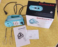 Shows the turquoise hand mixer, two metal beaters, original box with image of the mixer, and manual laying on a yellow surface.