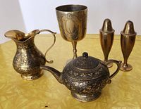 Five brass vintage pieces arranged on a yellow surface: teapot, creamer, salt & pepper shakers, and goblet