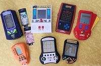 Top-down view of 8 vintage handheld games arranged on table showing various brands and designs including Pinball, Tactactoe, UNO, Armor Battle and others