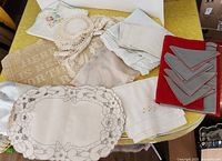 Lot items arranged on a yellow table: crocheted doilies, embroidered table linens and folded cloth napkins in beige, white, and red with grey trim.