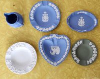 Top-down photo of six Wedgwood jasperware items on yellow fabric: four ashtrays, one small pitcher, and one small tray in shades of blue, white, and green with white classical relief decorations.