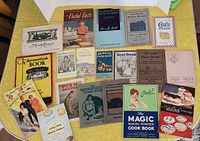 Top view of 20 vintage and antique cookbooks and recipe booklets laid out on yellow surface, covers visible showing titles and artwork circa 1920s and 1930s.