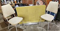 Two cream floral patterned chairs positioned on either side of a yellow table top with metal rails, photographed in a garage setting.