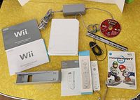 Full overview photo of Nintendo Wii console, controllers, manuals, stand, power adapter, sensor bar, and two Wii games including Family Feud and Mario Kart box with booklet only.