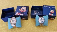 Pair of two Disney Mickey Mouse wristwatches displayed on blue pillows with original blue Disney boxes showing Mickey Mouse artwork. One watch is gold-tone with rhinestones, the other silver-tone metal.