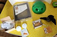 Top view of all items on yellow surface: PetSafe pet screen door, green Pop & Play dome toy, black flapping bat toy, Smart Bell dog training kit with two white bells, box of bully sticks with circular white disks on box